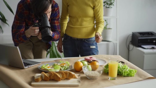 Female flatlay stylist arranging objects on table helping young male photographer to take food images in office