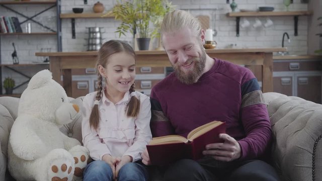Portrait of surprised Caucasian girl listening to father reading her book. Positive man touching daughter's nose with hand. Family having fun at home. Parenthood, hobby, lifestyle.
