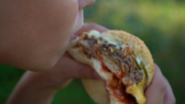 Closeup view of hungry kid eating big double cheeseburger standing outdoor. Macro of child mouth biting big meat burger. Slow motion full hd video footage.