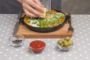 Stewed eggs and vegetables in a pan, shakshuka for breakfast. Hand grate cheese. Cheese, grater, olives, peppers on the table..