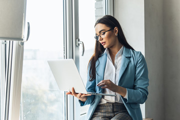 Young beautiful businesswoman in glasses working on laptop and keeping hand on chin while sitting...