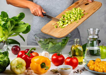 Woman adds chopped avocado to salad in a glass bowl, cooking in the kitchen. Healthy diet concept.