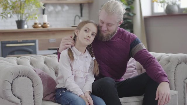Portrait of happy Caucasian father caressing daughter's head, looking at camera and smiling. Young man and teenage girl with braided pigtails posing at home. Parenthood, happiness, lifestyle.