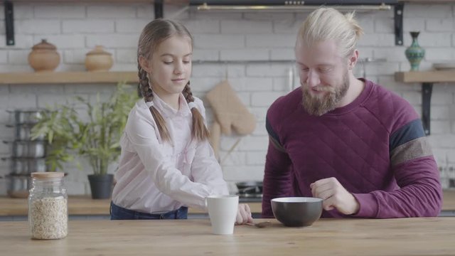Portrait of pretty Caucasian teenage girl giving father plate and spoon and opening bottle with corn flakes. Daughter taking care of adult bearded man. Happy amicable family in the kitchen at home.