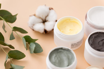 Fototapeta na wymiar beauty, spa and wellness concept - blue clay mask, body butter, therapeutic mud and eucalyptus cinerea with cotton flower on beige background