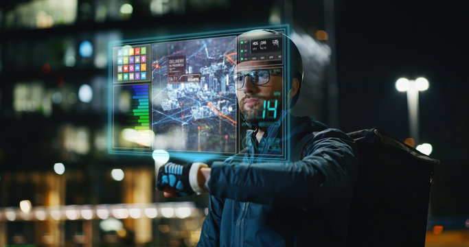 Portrait of an young delivery courier with bicycle is touching a futuristic screen with augmented reality hologram maps to view the addresses of customers in the evening in a city center.