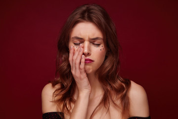 Studio shot of upset unhappy woman with brown wavy hair and festive makeup holding palm on her face and keeping eyes closed, almost going to cry while standing over claret background