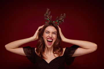 Overjoyed pretty young brunette woman with wavy hairstyle dressed in elegant clothes rejoicing while posing against claret background, wearing funny holiday hoop and laughing happily