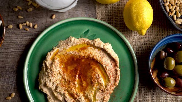Sliding to the left showing a flat lay plate of hummus with feta, olives, glass of ouzo, lemons, pine nuts on a rustic wood table.