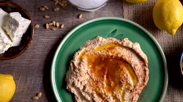 Sliding to the right showing a flat lay plate of hummus with feta, olives, glass of ouzo, lemons, pine nuts on a rustic wood table.