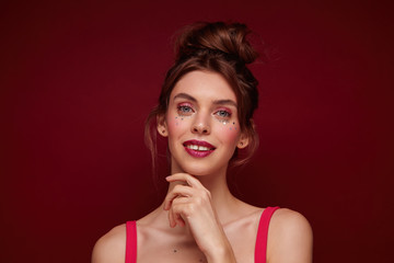 Studio shot of charming young brown haired female with bun hairstyle wearing festive makeup while posing over burgundy background, looking positively at camera and touching face gently