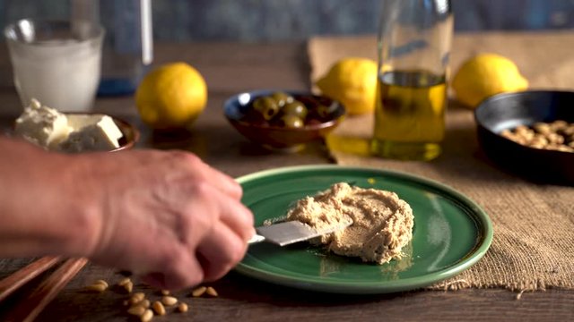 Beautiful setting of middle eastern food as hand comes in a spreads hummus onto a plate, pours olive oil and sprinkles paprika.