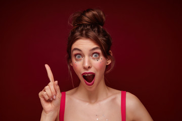 Close-up of shocked young pretty brown haired woman with casual hairstyle raising up index finger and looking at camera with wide eyes and mouth opened, isolated over burgundy background