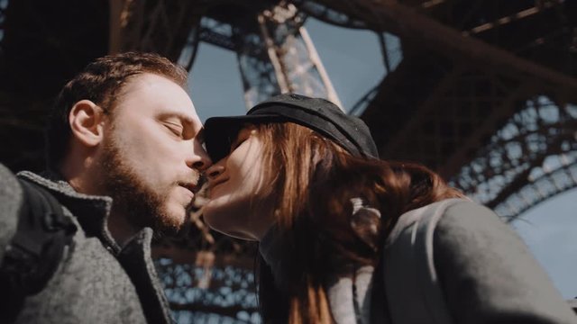 Close-up low angle happy smiling romantic couple kissing, looking at each other under Paris Eiffel Tower slow motion.