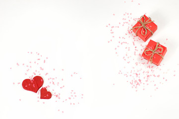 two red gift boxes in the upper right corner of the image and two red wooden hearts in the lower left corner are sprinkled with small shiny red hearts on a white background top view