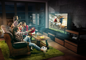 Group of friends watching TV, football match, sport together. Emotional men and women cheering for...