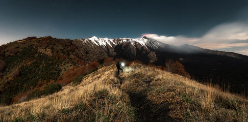 Man illuminating the mountains landscape with a torch. Volcano Etna covered by snow erupting in the night with full moon. Hiking in the mountains