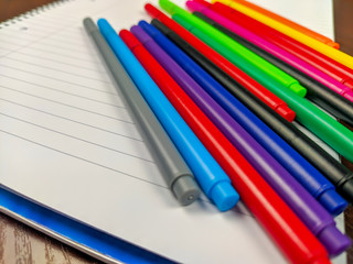Fototapeta premium Closeup of colorful pens laying on an empty white notebook for writing down notes as an equipment or supplies for school or studying
