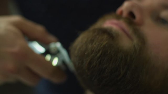 Bearded man and barbers hands shaving beard by electric shaving machine and comb in slow motion. Male with closed eyes in barbershop. 