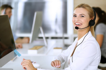 Blonde business woman using headset for communication and consulting people at customer service office. Call center. Group of operators at work at the background.