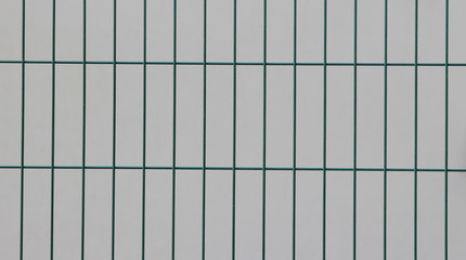 Surface of a welded wire fence with rectangular elements painted in green in front of a white wall 