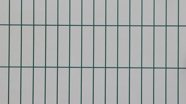 Surface of a welded wire fence with rectangular elements painted in green in front of a white wall 