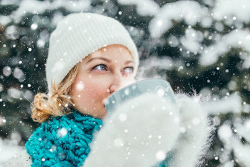 Young attractive blonde in white warm mittens, a scarf and a hat with a cup of hot drink in the forest - a nice New Year and Christmas look