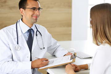 Doctor and patient discussing the results of a physical examination while sitting at a desk in a clinic. A male doctor using a clipboard to fill out a medical history of a young woman's medication