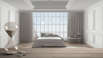 Wooden table, desk or shelf with crystal modern hourglass measuring the passing time and house keys over classic bedroom with master bed, architecture interior design, blur background