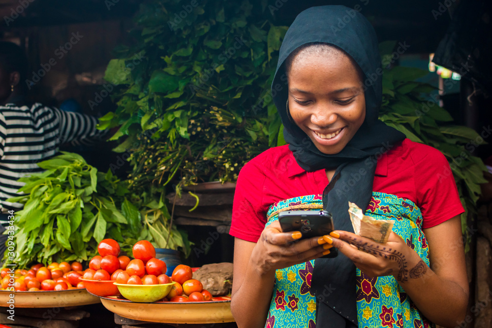 Wall mural young african woman selling in a local market smiling while using her mobile phone