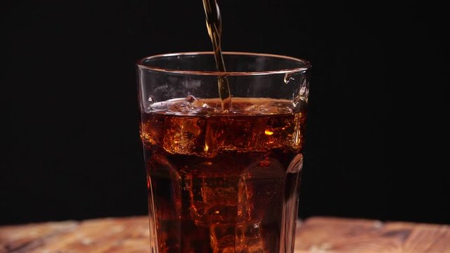Pouring of tasty cola into glass with ice on black background. Slow motion effect