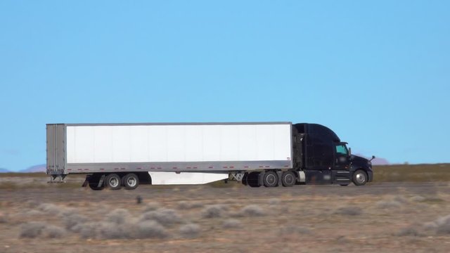 CLOSE UP: Black semi-trailer truck speeds along the interstate highway crossing a desert in United States. Cargo lorry hauls heavy freight container across the countryside of United States of America.