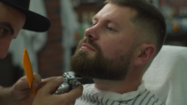 Bearded man and barbers hands shaving beard by electric shaving machine and comb in slow motion. Male with closed eyes in barbershop. 