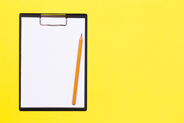 Clipboard with white sheet and pen on a yellow background. View from above. space for text