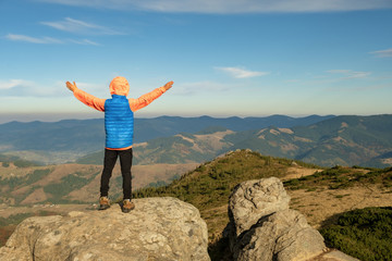 Young child boy hiker standing with raised hands in mountains enjoying view of amazing mountain landscape at sunset.