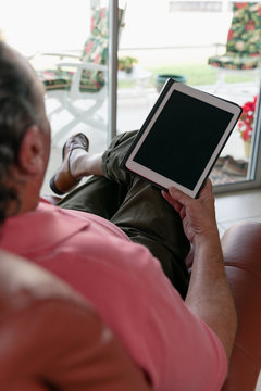 Over The Shoulder Of A Senior Man In His Seventies Holding A Tablet.