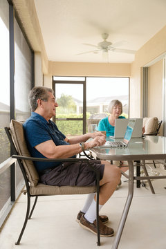 Senior couple in their seventies relaxes on their patio lanai while using their laptops.