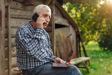 Elderly man with a laptop in village, in nature. Old man out of town with computer. Grandfather in...