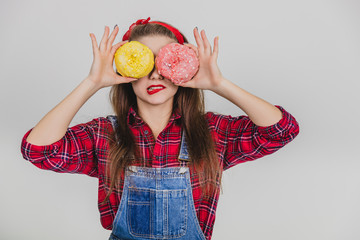 Lovely young female hiding her eyes behind two big yellow and pink doughnuts, twisting her lip.