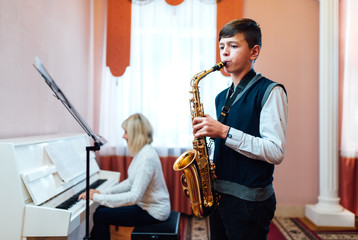 A teenage boy learns to play saxophone in a music lesson to accompaniment of a female teacher on the piano.