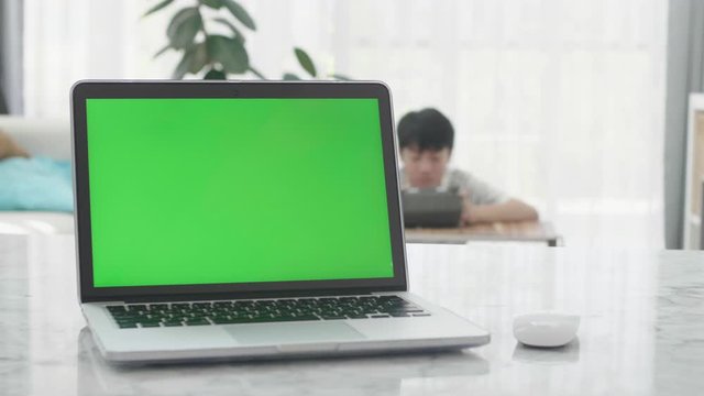 Laptop Computer Showing Green Chroma Key Screen Stands on a Desk in the Living Room. In the Background mother and son watching on tablet computer together.