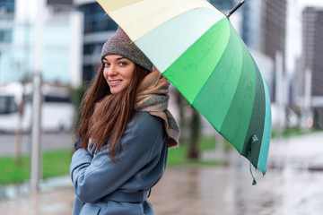 Portrait of happy smiling joyful carefree woman in warm clothes with bright multicolored rainbow umbrella during rainy day and rain weather in the autumn in the city. Rain protection