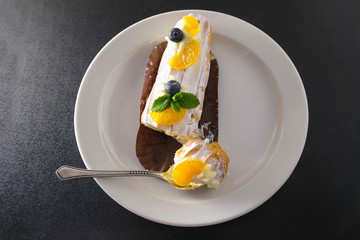 Cake with cream and fresh fruits on a white plate. Blueberry, tangerine slices and mint leaves. Piece of cake in a spoon.