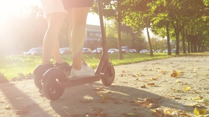 SLOW MOTION, LOW ANGEL, CLOSE UP, LENS FLARE: Two unrecognizable female friends ride e-scooters down gravel walkway. Millennial women ride high tech electric scooters down vivid avenue on a sunny day. - Powered by Adobe