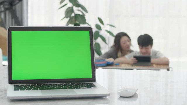 Laptop Computer Showing Green Chroma Key Screen Stands on a Desk in the Living Room. In the Background mother and son watching on tablet computer together.