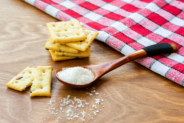 Crispy salted crackers, wooden spoon with salt crystalls, paper bag full of crackers on a red napkin on wooden table. Salty junk, unhealthy snacks, fast carbohydrates food.