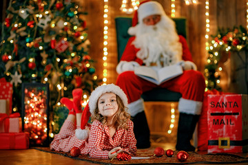 Little charming girl in pajama, Santa hat is lying on the floor and writes a letter, draws with a pencil. lights of garlands. warm comfortable room