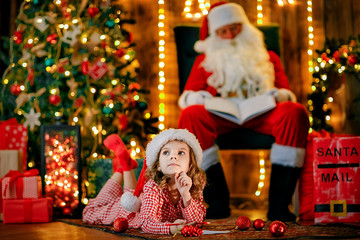 Little charming girl in pajama, Santa hat is lying on the floor and writes a letter, draws with a pencil. lights of garlands. warm comfortable room