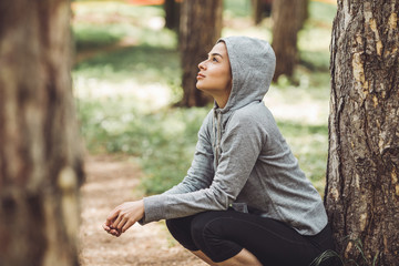 A young woman resting from jogging in the park by a tree. She is wearing a grey hoodie and gazes...