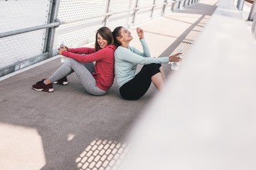 Two women resting on a running track on the bridge. They were jogging, now they're sitting back to back, one of them is drinking water.